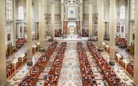 Wielka bazylika w Licheniu Nowym wypełniona wiernymi. Długie ławy zapełnione ludźmi, majestatyczne kolumny i ołtarz w tle.