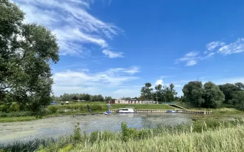Międzychód atrakcje: spokojna rzeka z łodziami przy brzegu, zielone drzewa i budynki w tle, błękitne niebo z chmurami.