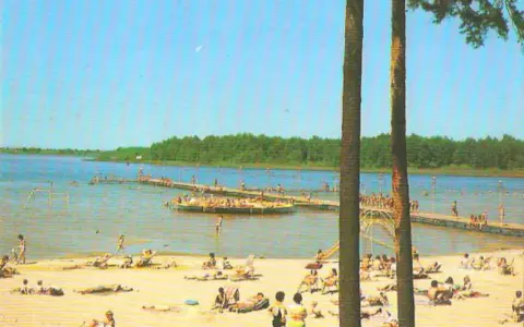 Słoneczny dzień nad jeziorem Niedzięgiel. Ludzie odpoczywają na plaży i pływają, a na wodzie unosi się platforma rekreacyjna.