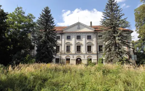 Dawny dwór otoczony przez wysokie trawy i drzewa, w tym sosny i dęby.