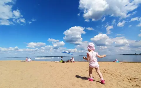 Dziewczynka w różowym kapelusiku bawi się na plaży przy zaporze Jeziorsko. W tle widać ludzi odpoczywających pod parasolem i spokojną taflę wody.