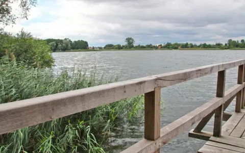 Drewniana kładka nad spokojnym jeziorem Lednica, otoczona trzcinami i zielenią.