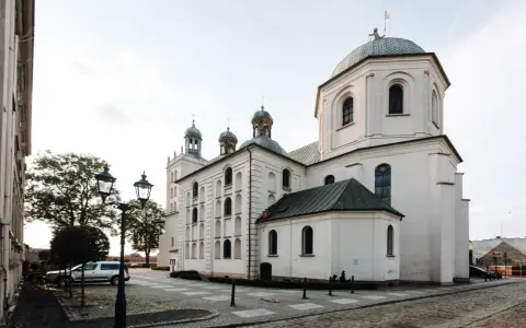 Majestatyczna fara w Grodzisku Wlkp. z charakterystycznymi kopułami i wieżami, otoczona brukowanym placem i zabytkowymi latarniami.