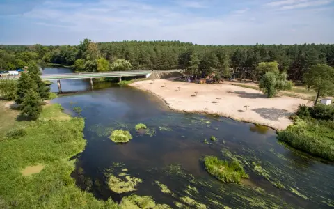 Malownicza plaża nad jedną z wielkopolskich rzek, z mostem i lasem w tle. Idealne miejsce na wypoczynek.