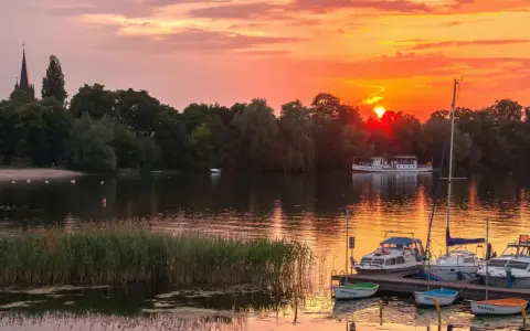Zachód słońca nad jeziorem z piaszczystą plażą w Polsce. Łodzie zacumowane przy pomoście, na wodzie pływają łabędzie.