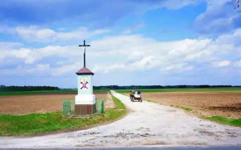 Krzyż przy drodze, obok pola i lasu. Na drodze widać wóz ciągnięty przez konie, z ludźmi.