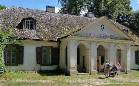 Stare dworki polskie z omszałym dachem i werandą. Rodzina z wózkiem spaceruje po dziedzińcu.