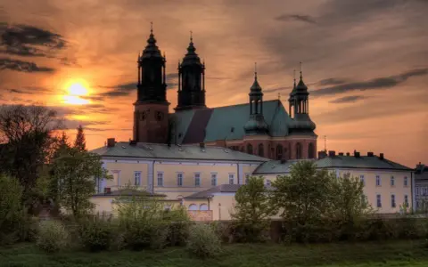 Zachód słońca nad katedrą, najstarszym kościołem w Polsce. Monumentalna budowla z cegły i zielonym dachem, otoczona drzewami.
