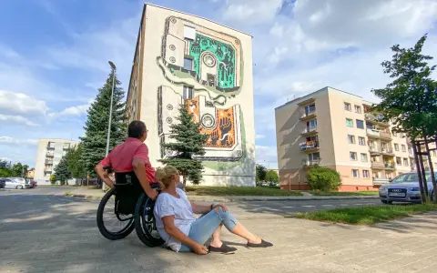 Para w Jarocinie podziwia mural na bloku. On na wózku, ona siedzi obok.