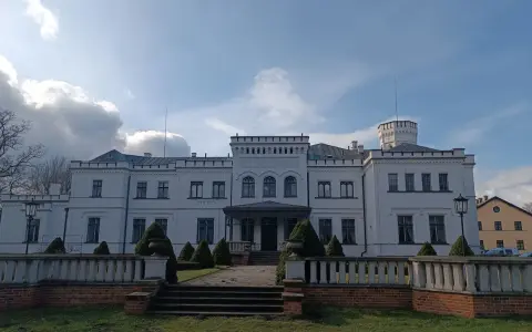 Biały pałac w Będlewie z wieżyczką i ozdobnym murem. Słońce przebija się przez chmury, tworząc malowniczy krajobraz.