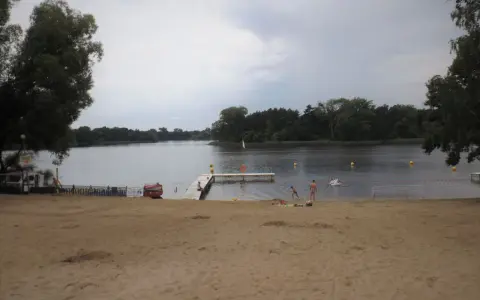 Plaża nad jeziorem koło Gniezna. Ludzie odpoczywają, niektórzy kąpią się. W tle las i spokojna woda.