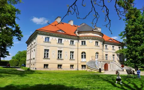 Pałac w Lewkowie, żółty, z pomarańczowym dachem, otoczony zielenią. Dzieci bawią się na trawniku przed budynkiem.