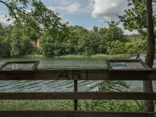 Widok na jezioro w Wielkopolskim Parku Narodowym. Na pierwszym planie drewniana balustrada z tablicami informacyjnymi. W oddali las i ruiny ceglanej budowli.