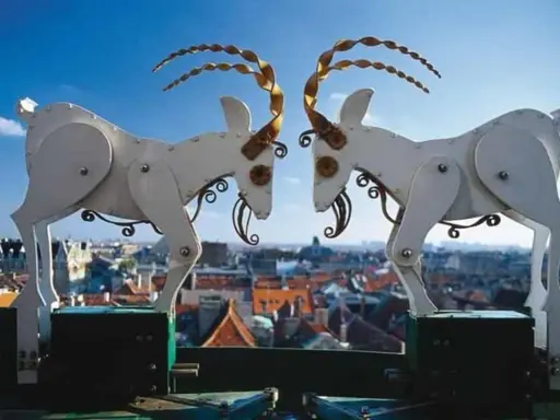 Mechaniczne koziołki poznańskie na tle miasta. Dwa białe, metalowe koziołki z kręconymi rogami stykają się głowami.