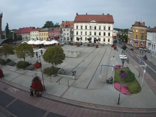 Rynek w Czarnkowie z charakterystycznym kościołem po lewej stronie. Plac tętni życiem, widać ludzi, fontanny i kawiarniane stoliki.