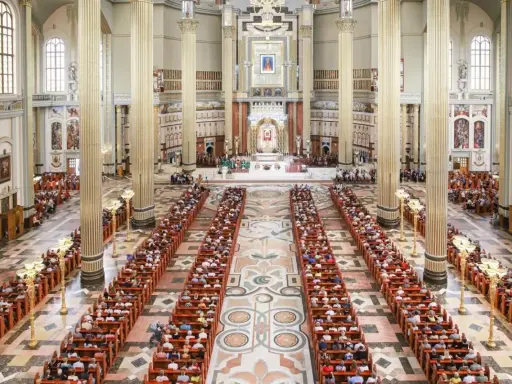 Wielka bazylika w Licheniu Nowym wypełniona wiernymi. Długie ławy zapełnione ludźmi, majestatyczne kolumny i ołtarz w tle.