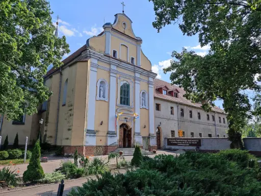 Żółty kościół w Szamotułach z białymi zdobieniami, otoczony zielenią. Obok tablica informująca o adoracji.