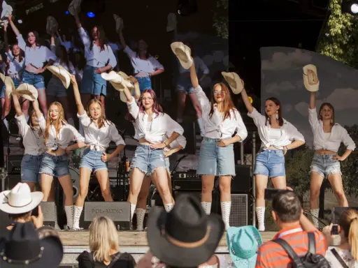 Dziewczęcy zespół country z Wolsztyna w białych koszulach i jeansowych szortach tańczy na scenie, unosząc kapelusze.