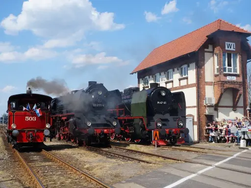 Parada parowozów z parą unoszącą się w powietrzu. Trzy lokomotywy stoją na torach obok ceglanego budynku stacji, a tłum widzów obserwuje wydarzenie.