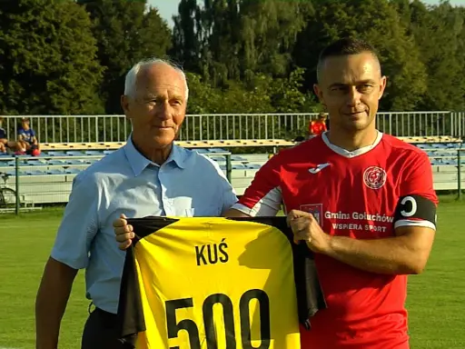 Dwóch mężczyzn na boisku, jeden w niebieskiej koszuli, drugi w czerwonej, trzymają żółtą koszulkę z napisem "KUŚ 500". To ważne wydarzenia Chodzież.