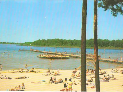 Słoneczny dzień nad jeziorem Niedzięgiel. Ludzie odpoczywają na plaży i pływają, a na wodzie unosi się platforma rekreacyjna.