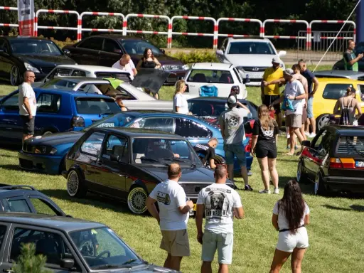 Zlot fanów motoryzacji w Łobżenicy. Wiele samochodów, ludzie oglądają auta, atmosfera piknikowa.