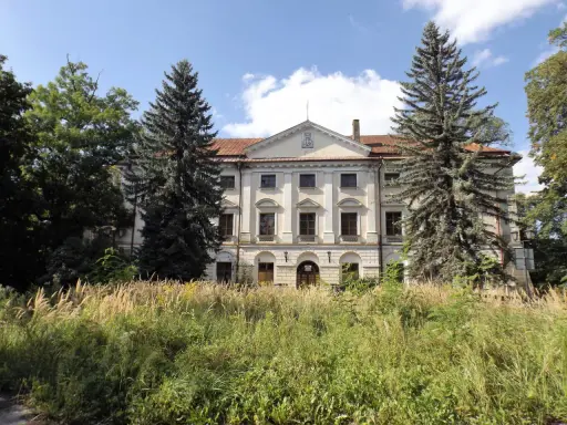 Dawny dwór otoczony przez wysokie trawy i drzewa, w tym sosny i dęby.