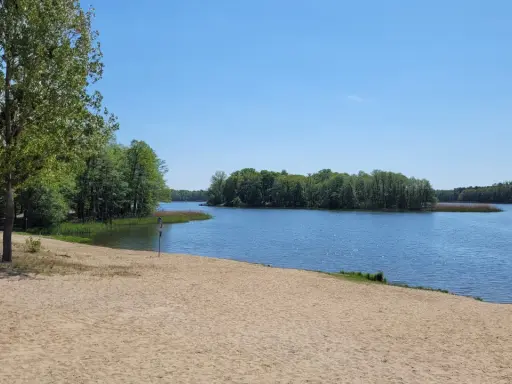 Piękny dzień nad jeziorem w Wielkopolsce. Piaszczysta plaża, spokojna woda i zielone drzewa tworzą idealne miejsce na wypoczynek.