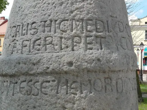 Kamienny słup milowy z wyrytymi łacińskimi napisami, stojący na tle budynków i zieleni.