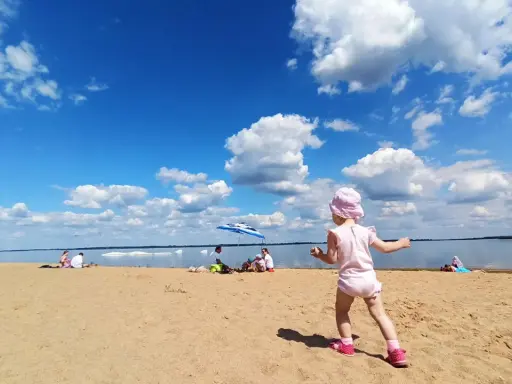 Dziewczynka w różowym kapelusiku bawi się na plaży przy zaporze Jeziorsko. W tle widać ludzi odpoczywających pod parasolem i spokojną taflę wody.