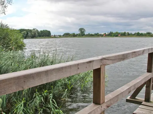 Drewniana kładka nad spokojnym jeziorem Lednica, otoczona trzcinami i zielenią.