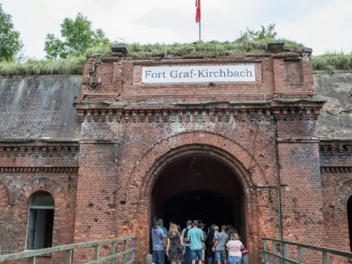 Wejście do Fortu Graf-Kirchbach, historycznej twierdzy w Poznaniu. Grupa turystów wchodzi do ciemnego tunelu.