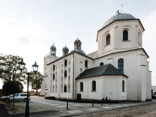 Majestatyczna fara w Grodzisku Wlkp. z charakterystycznymi kopułami i wieżami, otoczona brukowanym placem i zabytkowymi latarniami.