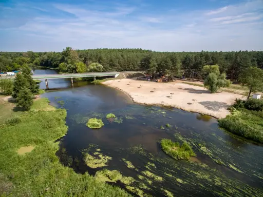 Malownicza plaża nad jedną z wielkopolskich rzek, z mostem i lasem w tle. Idealne miejsce na wypoczynek.