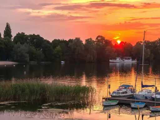 Zachód słońca nad jeziorem z piaszczystą plażą w Polsce. Łodzie zacumowane przy pomoście, na wodzie pływają łabędzie.