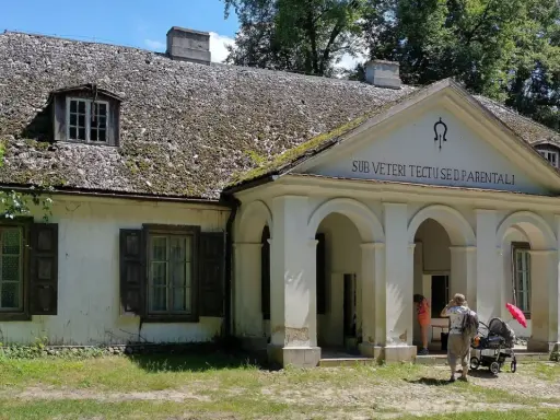 Stare dworki polskie z omszałym dachem i werandą. Rodzina z wózkiem spaceruje po dziedzińcu.