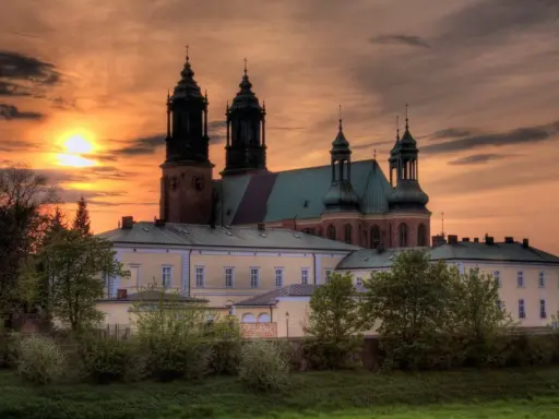 Zachód słońca nad katedrą, najstarszym kościołem w Polsce. Monumentalna budowla z cegły i zielonym dachem, otoczona drzewami.