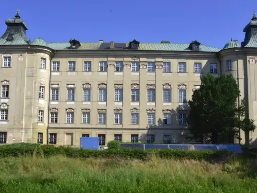 Imponujący pałac, największy zamek w Wielkopolsce, z zielonymi dachami i licznymi oknami, otoczony drzewami i trawą.
