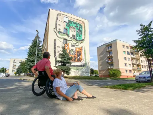 Para w Jarocinie podziwia mural na bloku. On na wózku, ona siedzi obok.