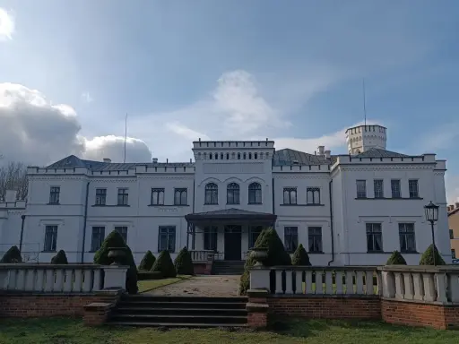 Biały pałac w Będlewie z wieżyczką i ozdobnym murem. Słońce przebija się przez chmury, tworząc malowniczy krajobraz.