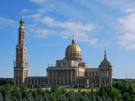 Imponujące sanktuarium koło Konina z wysoką wieżą i złotą kopułą, otoczone zielenią.