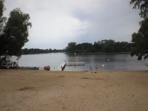 Plaża nad jeziorem koło Gniezna. Ludzie odpoczywają, niektórzy kąpią się. W tle las i spokojna woda.