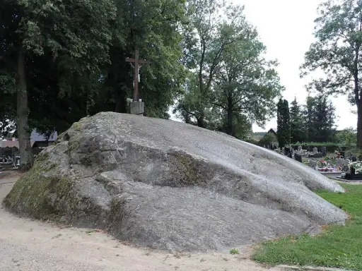 Ogromny głaz narzutowy, na którym stoi krzyż. W tle cmentarz i drzewa.
