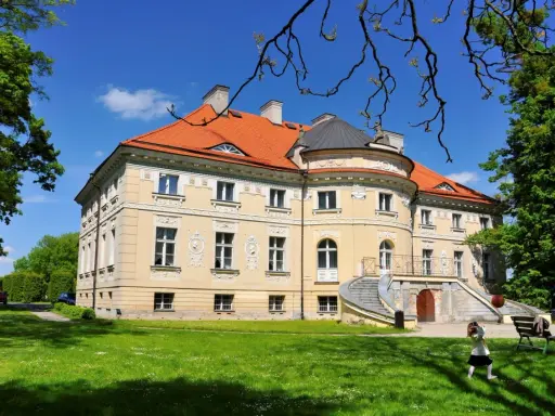 Pałac w Lewkowie, żółty, z pomarańczowym dachem, otoczony zielenią. Dzieci bawią się na trawniku przed budynkiem.