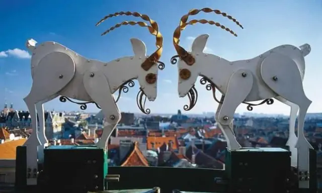 Mechaniczne koziołki poznańskie na tle miasta. Dwa białe, metalowe koziołki z kręconymi rogami stykają się głowami.