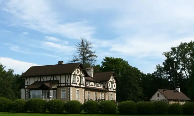 Elegancka zabudowa szachulcowa z ciemnym dachem i białymi ścianami, otoczona zielenią, pod błękitnym niebem.