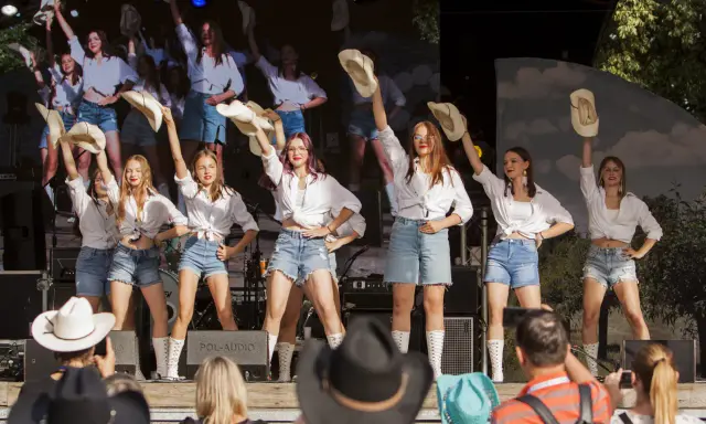 Dziewczęcy zespół country z Wolsztyna w białych koszulach i jeansowych szortach tańczy na scenie, unosząc kapelusze.