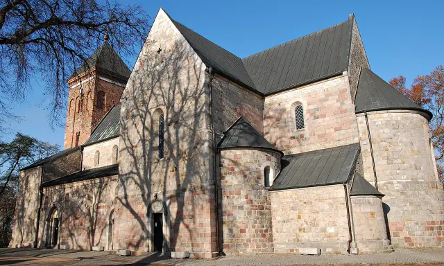Kamienna budowla z ceglaną wieżą, z cieniem drzew na ścianach, w stylu romańskim, przed gotykiem.