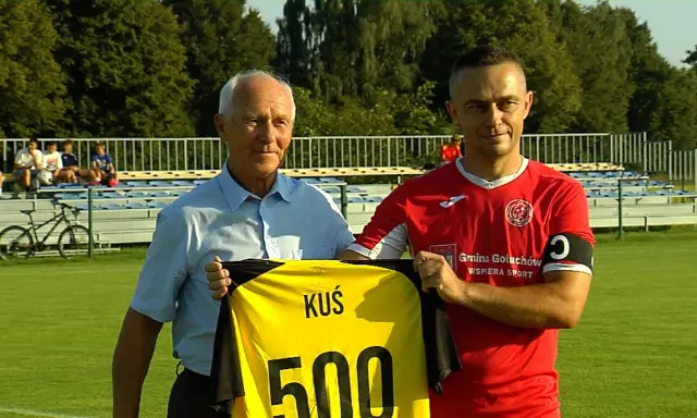 Dwóch mężczyzn na boisku, jeden w niebieskiej koszuli, drugi w czerwonej, trzymają żółtą koszulkę z napisem "KUŚ 500". To ważne wydarzenia Chodzież.