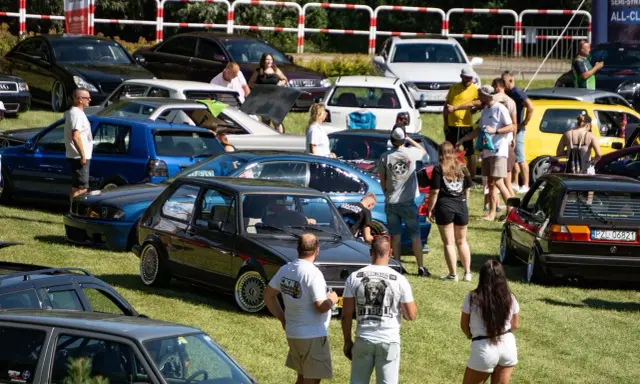 Zlot fanów motoryzacji w Łobżenicy. Wiele samochodów, ludzie oglądają auta, atmosfera piknikowa.